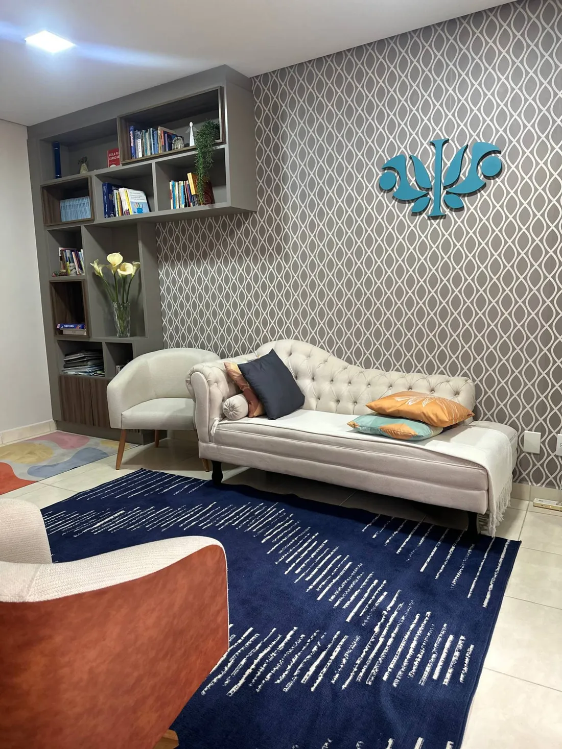 Divã capitonê com almofadas coloridas, estante de livros e flores no consultório